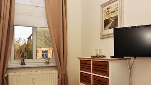 Une pièce avec une fenêtre, un radiateur, une armoire en bois, un téléviseur et un tableau encadré au mur.
