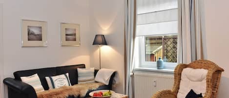 Un salon avec un canapé noir, un fauteuil en osier, une lampe et une fenêtre avec des rideaux.