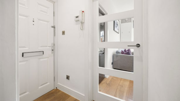 A white door with a phone, a mirror, and a view into a room with a gray sofa and purple pillows. - Marylebone’s Nest 1Bed (London)