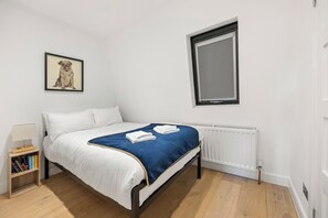 A bedroom with a bed, a radiator, a window, a framed picture on the wall, and a nightstand with books. - Marylebone’s Nest 1Bed (London)