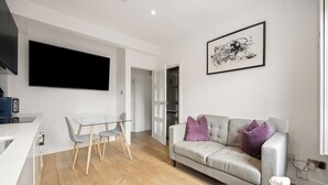 A modern living room with a flat-screen TV, a grey sofa with purple cushions, a glass dining table with chairs, and a white armchair. - Marylebone’s Nest 1Bed (London)