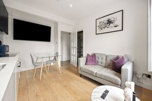 A modern living room with a flat-screen TV, a grey sofa with purple cushions, a glass dining table with chairs, and a white armchair. - Marylebone’s Nest 1Bed (London)