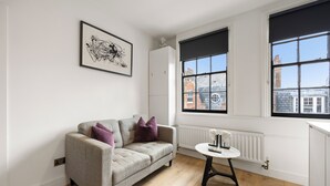 A modern living room with a sofa, a small table, and large windows with black blinds. - Marylebone’s Nest 1Bed (London)