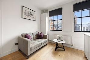 A modern living room with a sofa, a small table, and large windows with black blinds. - Marylebone’s Nest 1Bed (London)