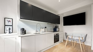 A modern kitchen with white cabinetry, a black range hood, a built-in oven, and a glass dining table with two chairs. - Marylebone’s Nest 1Bed (London)