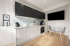 A modern kitchen with white cabinetry, a black range hood, a built-in oven, and a glass dining table with two chairs. - Marylebone’s Nest 1Bed (London)