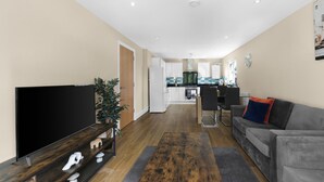 A modern living room with a flat-screen TV, a wooden coffee table, a grey sofa, and a kitchen in the background. - Heathrow Living 2BDR 2BA Apt by 360 Stays - Apt 12 (Stanwell)
