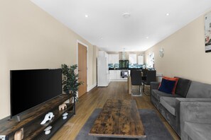 Ein modernes Wohnzimmer mit Flachbildfernseher, einem Holztisch, einer grauen Couch und einer Küche im Hintergrund.