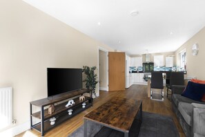 Ein modernes Wohnzimmer mit Flachbildfernseher, Couchtisch und Sofa. Im Hintergrund ist eine Küche mit weißen Schränken und einem Essbereich zu sehen.