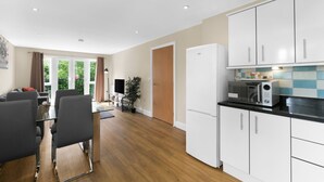 A modern kitchen with white cabinets, a microwave, and a refrigerator. There is a dining area with a table and chairs, and a large window with curtains. - Heathrow Living 2BDR 2BA Apt by 360 Stays - Apt 12 (Stanwell)