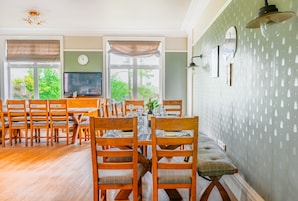 Un comedor con sillas y mesas de madera, un televisor y vistas al exterior con vegetación.