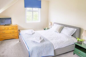 Un dormitorio con una cama, un televisor sobre un aparador de madera, una ventana con persiana azul y una mesita de noche con una lámpara.