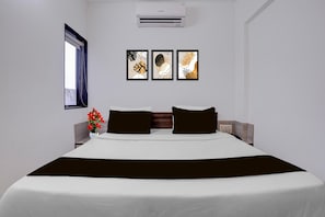A hotel room with a bed, a window, an air conditioner, and three framed artworks on the wall.