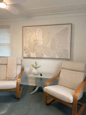 A room with two beige armchairs, a small white table with a vase and a remote control, and a large leaf-patterned artwork on the wall. - NEW! Restored Oceanfront Cottage •  Vintage Charm • Walk to Dining (Topsail Beach)