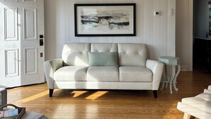 A living room with a white leather sofa, a framed abstract painting, and a wooden floor. - NEW! Restored Oceanfront Cottage •  Vintage Charm • Walk to Dining (Topsail Beach)