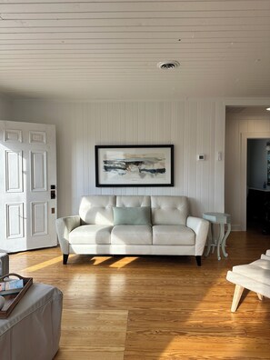 A living room with a white leather sofa, a framed abstract painting, and a wooden floor. - NEW! Restored Oceanfront Cottage •  Vintage Charm • Walk to Dining (Topsail Beach)