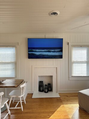 A room with a fireplace, a television mounted above it, and a view of the ocean through the window. - NEW! Restored Oceanfront Cottage •  Vintage Charm • Walk to Dining (Topsail Beach)