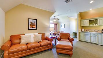 A living room with a brown leather sofa and armchair, a dining table with chairs, and a kitchen area with light green cabinets.