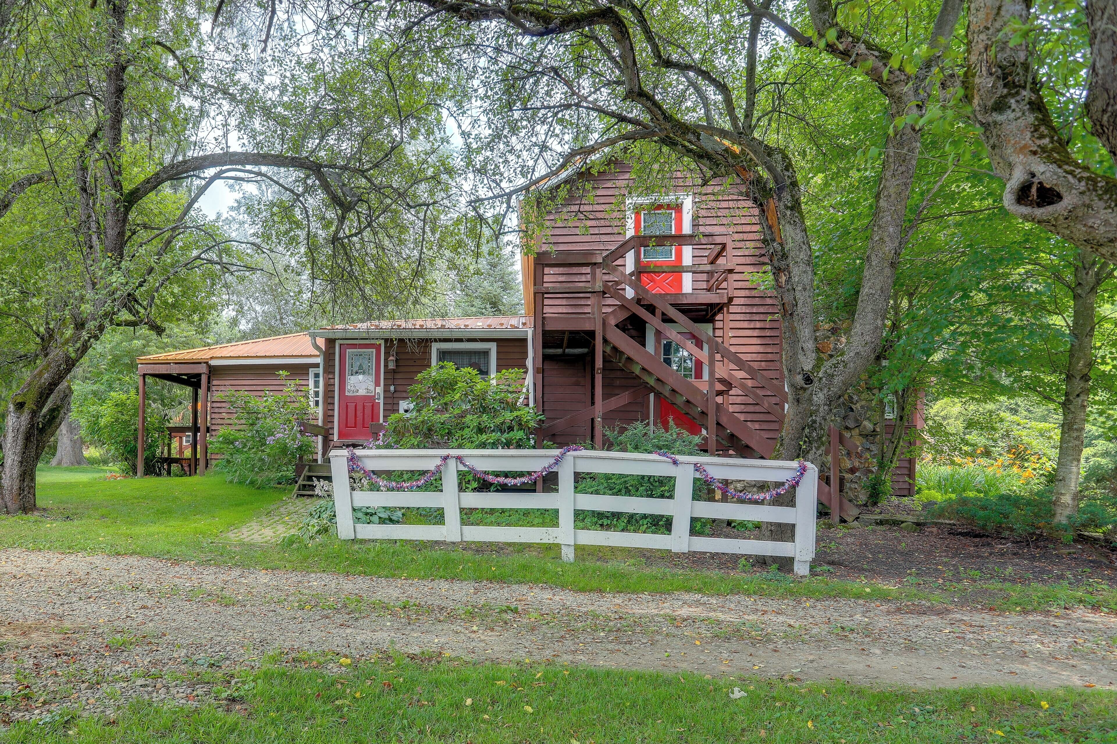A rustic cabin with a wooden staircase, surrounded by lush greenery and a white fence.