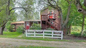 A rustic cabin with a wooden staircase, surrounded by lush greenery and a white fence.