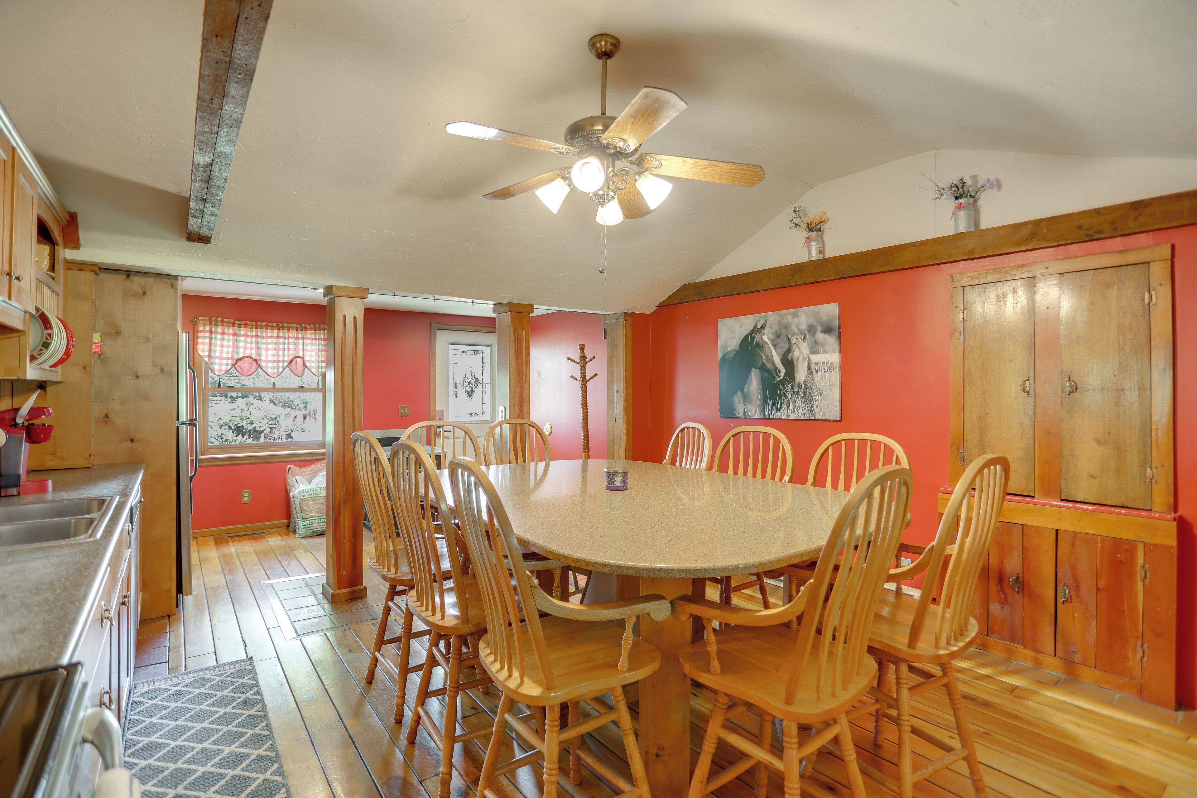 A dining area with a round table and wooden chairs, a ceiling fan, and a red wall with a painting.