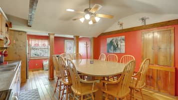 A dining area with a round table and wooden chairs, a ceiling fan, and a red wall with a painting.