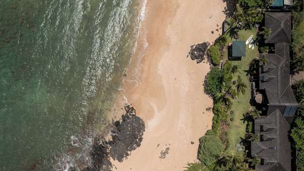 Luchtbeeld van een landgoed aan de kust met strand, rotsachtige kustlijn en een huis met schuin dak.