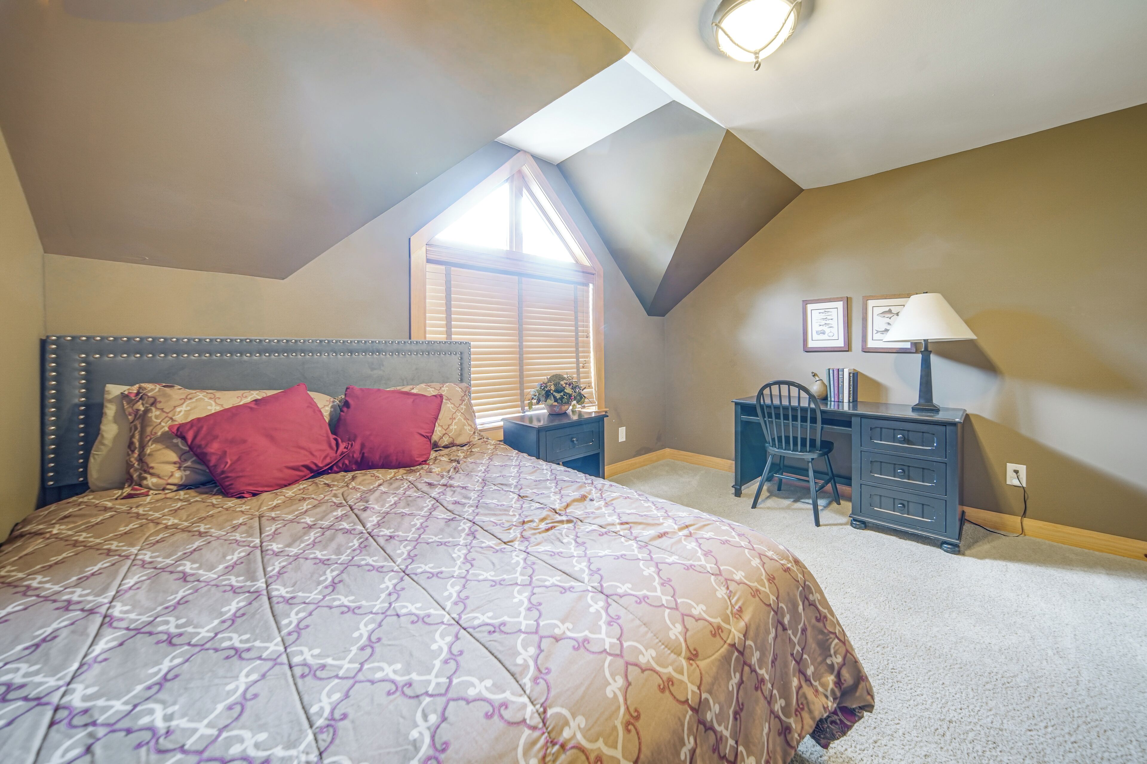 A bedroom with a sloped ceiling, a bed with patterned bedding, a desk with a lamp, and a window with blinds.