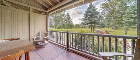 Une véranda couverte avec une table et des chaises en bois, une balustrade et une vue sur un espace verdoyant bordé d’arbres.