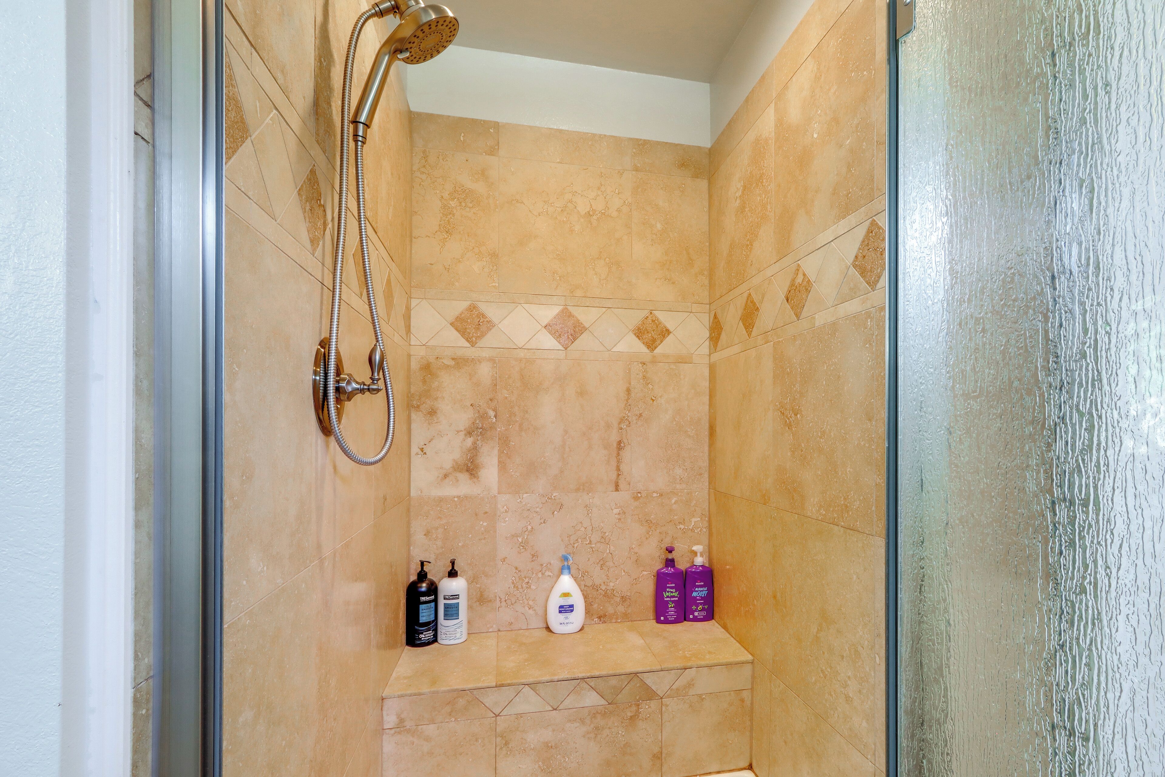 A shower with a handheld showerhead, a glass door, and a shelf with toiletries.
