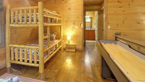 Interior de una cabaña de madera con literas, una mesita con una lámpara y una mesa de truco.