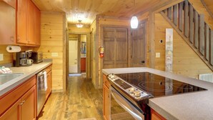 Interior de una cabaña de madera con una zona de cocina que incluye estufa, armarios y una escalera de madera.