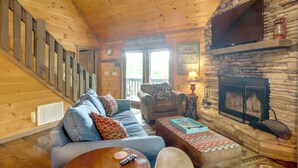 Interior acogedor de una cabaña con escalera de madera, chimenea, sofá y televisor.