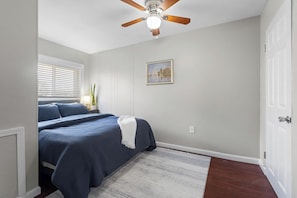 Ein Schlafzimmer mit Bett, Deckenventilator, Fenster mit Jalousien, einem Bild an der Wand und einer Tür.