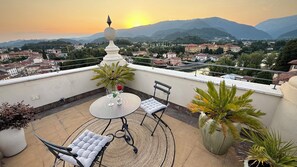 A rooftop terrace with a table and chairs, potted plants, and a view of a town and mountains. - Ponte degli Alpini | Luxury Penthouse · Sauna, 2 Hot Tubs & Panoramic Terrace (Bassano del Grappa)