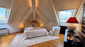 Ein Schlafzimmer mit schrägen Wänden, einem Bett mit weißen Leinen, Nachttischlampen, einer roten Lampe auf einem Nachttisch und einem Ausblick durch ein Fenster.