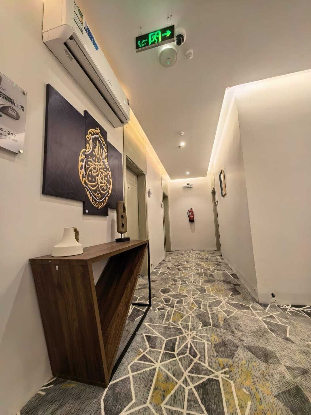 A hallway with a patterned carpet, a wall-mounted air conditioner, a decorative wall piece, and an exit sign.