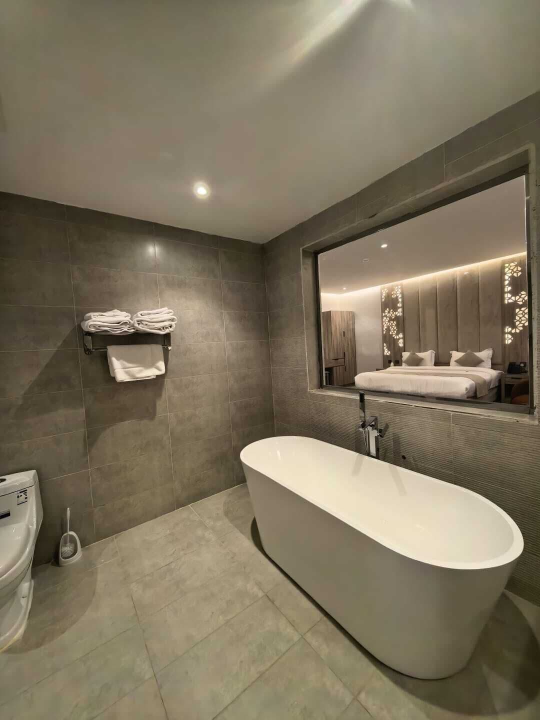 A modern bathroom with a freestanding bathtub, a toilet, and a large mirror reflecting a bedroom with a bed and nightstands.