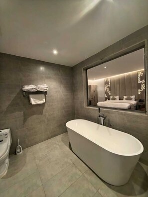 A modern bathroom with a freestanding bathtub, a toilet, and a large mirror reflecting a bedroom with a bed and nightstands. - Dar Al Eshaal Hotel (Madinah)