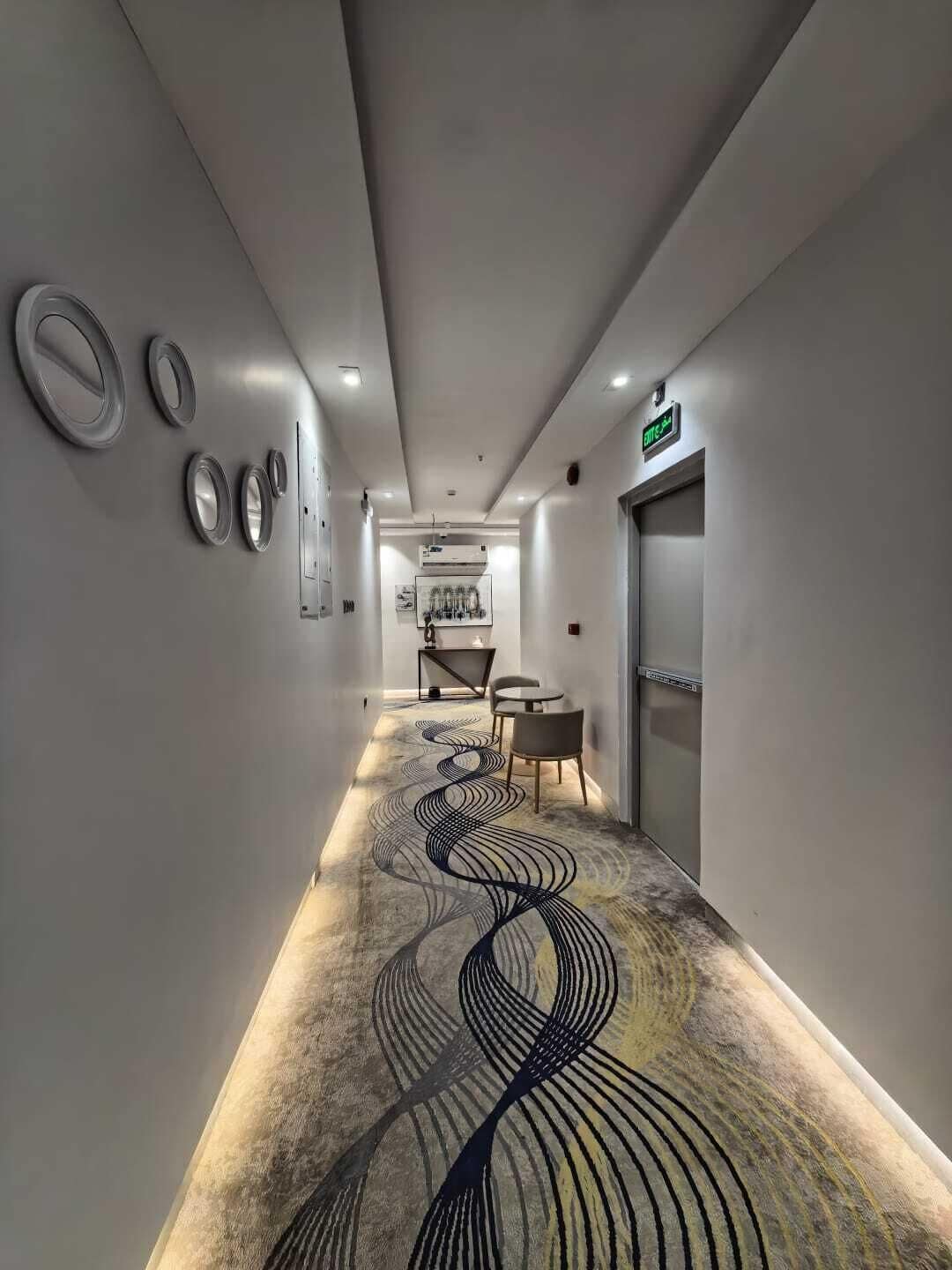 Un pasillo con alfombra estampada, una mesa con sillas y una puerta con señal de salida de emergencia.