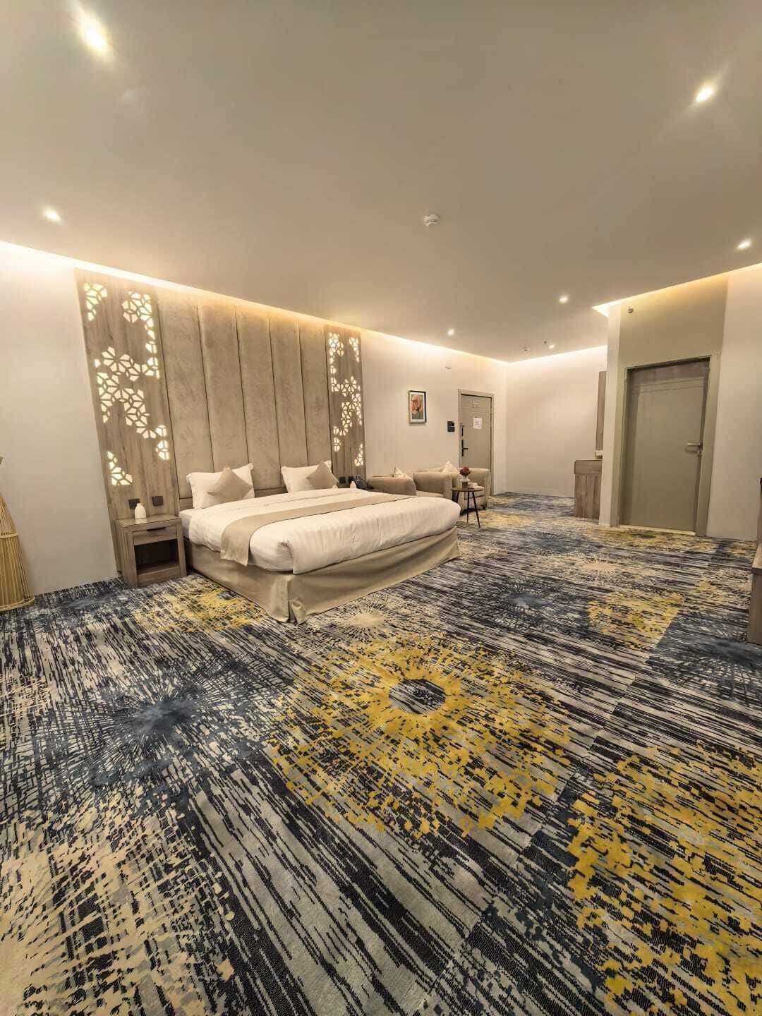 A spacious hotel room with a large bed, a small seating area, and a patterned carpet.