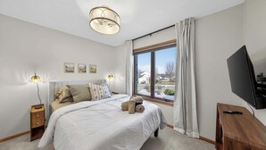 Een slaapkamer met een bed, nachtlampjes, een televisie, een raam met gordijnen en uitzicht op het omliggende landschap.