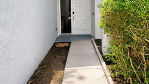 Un mur extérieur blanc surmonté d’un toit bleu, un chemin de béton menant à une porte blanche et un arbuste vert sur la droite.