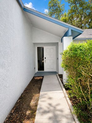 A white exterior wall with a blue roof, a concrete pathway leading to a white door, and a green bush to the right. - Cozy Tampa Home (Tampa)