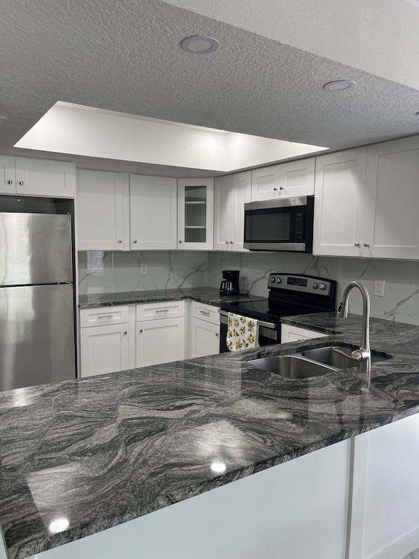 A modern kitchen with white cabinets, a stainless steel refrigerator, a microwave, a stove, and a granite countertop. - Cozy Tampa Home 2BR, 2BA & Fenced Yard (Tampa)