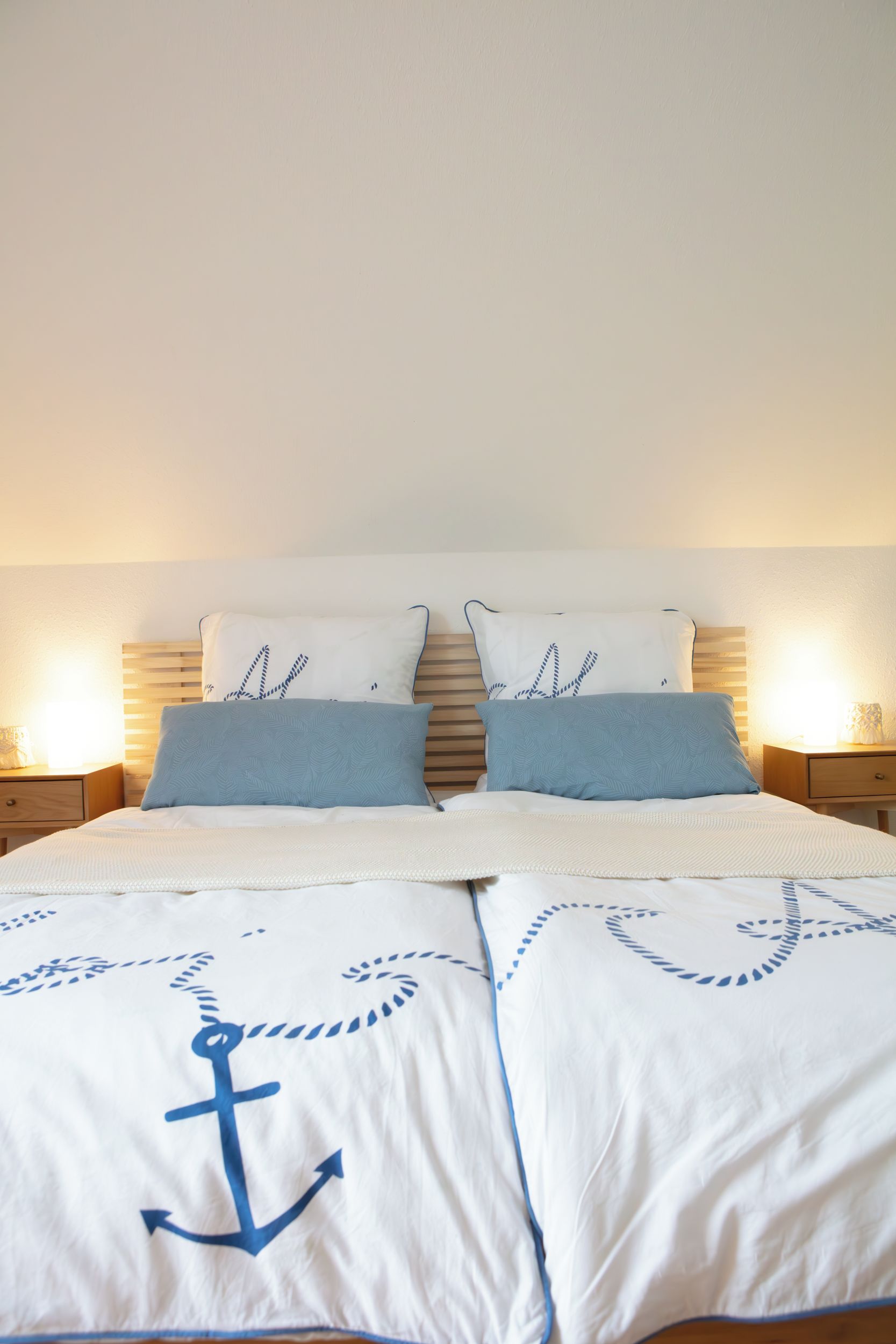 Una cama con cabecera de madera, dos almohadas con estampado de hojas y una colcha con diseño de ancla náutica.