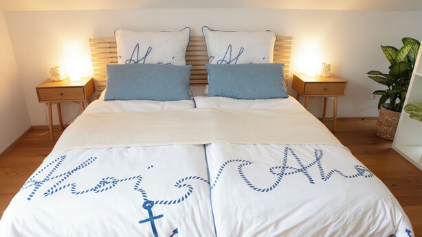 Un lit recouvert d’une courtepointe blanche ornée d’un motif d’ancre marine, flanqué de deux coussins assortis et de deux tables de chevet avec des lampes.