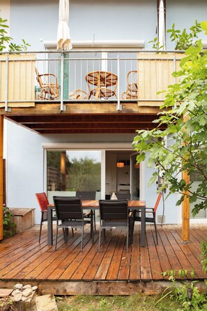 Eine Holzterrasse mit Esstisch und Stühlen, ein überdachter Sitzbereich mit Korbstühlen und Blick ins Haus.