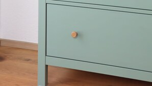 Une commode en bois vert clair, dotée de trois tiroirs et d’un plateau en bois, est placée contre un mur blanc.