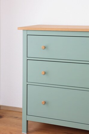 Eine hellgrüne Holzkommode mit drei Schubladen und einer Holzbalkonplatte vor einer weißen Wand.
